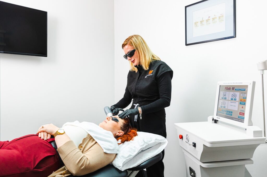 Wide shot of a patient lying on a treatment table and a provider using a microneedling device on her face. Before her appointment, she had a consultation to explore the differences between microdermabrasion vs. microneedling.